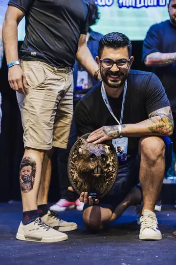 George Winck segurando o troféu da categoria retrato ao lado do cliente com o retrato de bebê tatuado na perna